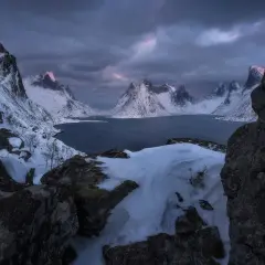 Foto panoramica de montañas nevadas y un lago oscuro bajo un cielo tormentoso. La luz del amanecer tiñe de rosa las cumbres.