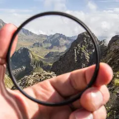 Mano sostiene un filtro UV para que sirve, mostrando un paisaje montañoso brumoso.