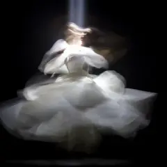 Figura etérea con un vestido blanco vaporoso, capturada con un **efecto foto movida** que evoca un baile onírico bajo un haz de luz.