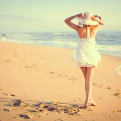 Mujer con sombrero blanco y vestido veraniego en la playa, disfrutando del mar. Estas fotos originales capturan la esencia del verano.