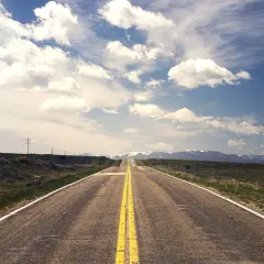 Carretera recta con doble línea amarilla que se pierde en el horizonte, flanqueada por campos verdes y montañas nevadas.