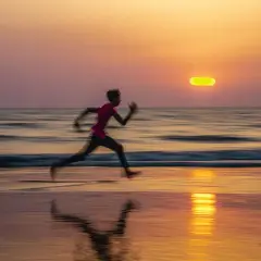 Persona corriendo en la playa al atardecer, con el sol anaranjado y reflejos en el agua. Foto barrido que captura el movimiento.