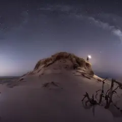 Persona ilumina la noche estrellada con su linterna, cerca de una tienda de campaña en la playa. Las fotos con ruido capturan la magia del cielo nocturno.