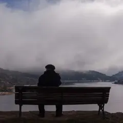 Hombre sentado en un banco, contemplando un lago y montañas cubiertas de niebla. La figura y el fondo crean una escena de introspección.