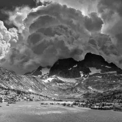 Paisaje montañoso dramático con nubes imponentes, típico de las ansel adams fotos. Un lago sereno refleja las cumbres nevadas.
