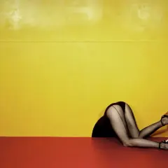 Piernas con medias de rejilla y tacones, estilo Guy Bourdin, asomando por un hueco contra un fondo amarillo y rojo.