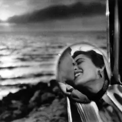 Una mujer sonríe en el espejo retrovisor de un coche, reflejando un beso. Elliott Erwitt capturó este momento íntimo junto al mar.