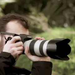Hombre con gafas y barba toma una foto con una cámara Canon. El parasol de la lente protege de la luz.
