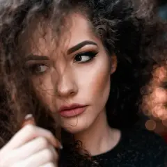 Plano medio corto de una mujer con cabello rizado y maquillaje dramático, mirando hacia un lado con una expresión pensativa.