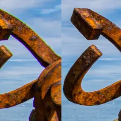 Escultura de metal oxidado con aberración cromática visible en los bordes, contra un cielo azul y mar.