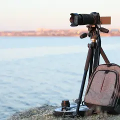 Equipo de fotografia basica listo para capturar el paisaje: cámara en trípode, mochila y lente junto al mar.