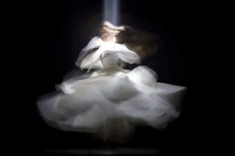 Figura etérea con un vestido blanco vaporoso, capturada con un **efecto foto movida** que evoca un baile onírico bajo un haz de luz.