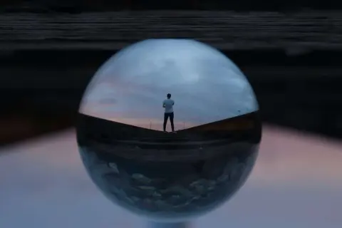 Un hombre mira el atardecer a través de una esfera de cristal, creando fotos originales y creativas.
