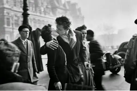 Pareja besándose apasionadamente en París, rodeada de gente y un coche antiguo. Un momento capturado en estas fotos históricas.