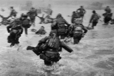 Soldados avanzan en el agua durante el desembarco de Normandía, una escena icónica capturada por Robert Capa.