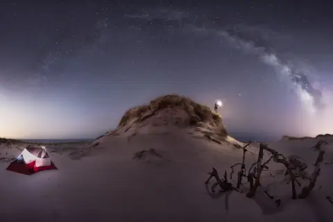 Persona ilumina la noche estrellada con su linterna, cerca de una tienda de campaña en la playa. Las fotos con ruido capturan la magia del cielo nocturno.