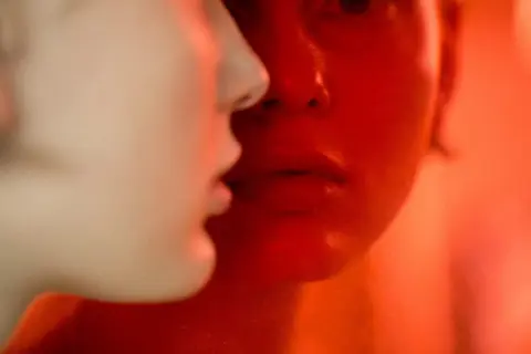 Primer plano de dos rostros, uno iluminado en rojo intenso, el otro en tonos claros. Martina Matencio captura un momento íntimo y enigmático.