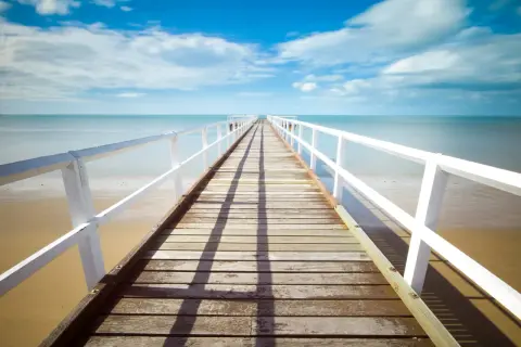 Un muelle de madera se extiende hacia el mar azul bajo un cielo nublado. La foto perspectiva crea una sensación de profundidad y calma.