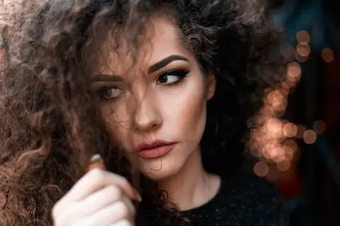 Plano medio corto de una mujer con cabello rizado y maquillaje dramático, mirando hacia un lado con una expresión pensativa.