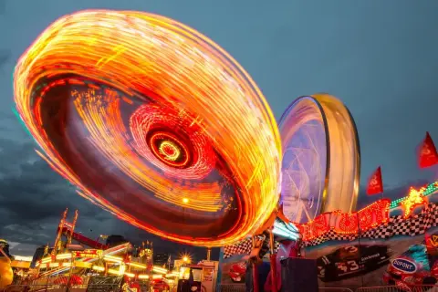 Luces de feria en movimiento capturadas con **fotos larga exposición**. Un torbellino de colores naranjas y rojos ilumina la noche, mientras una noria se vislumbra al fondo.