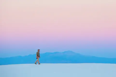 Hombre camina sobre dunas blancas bajo un cielo degradado de rosa y azul, respetando la ley del horizonte.