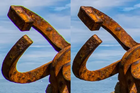Escultura de metal oxidado con aberración cromática visible en los bordes, contra un cielo azul y mar.