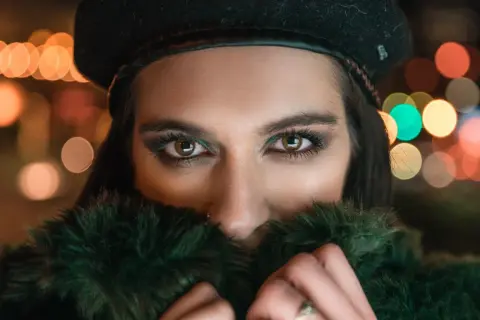 Ojos penetrantes de mujer con boina negra y abrigo de pelo verde, rodeada de luces bokeh. Un reto fotográfico nocturno.
