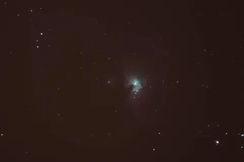 Nebulosa verdosa en el cielo nocturno, rodeada de estrellas y con tenues estelas de estrellas.