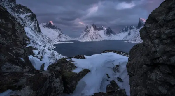 Foto panoramica de montañas nevadas y un lago oscuro bajo un cielo tormentoso. La luz del amanecer tiñe de rosa las cumbres.
