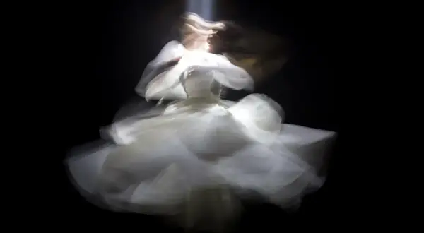 Figura etérea con un vestido blanco vaporoso, capturada con un **efecto foto movida** que evoca un baile onírico bajo un haz de luz.