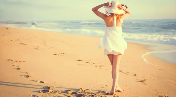 Mujer con sombrero blanco y vestido veraniego en la playa, disfrutando del mar. Estas fotos originales capturan la esencia del verano.