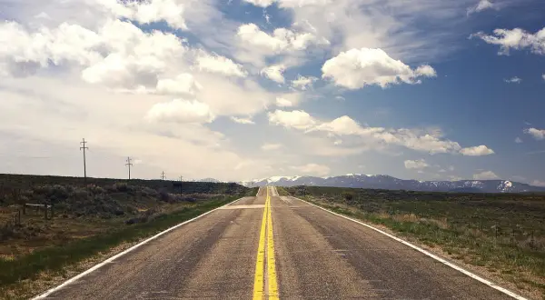 Carretera recta con doble línea amarilla que se pierde en el horizonte, flanqueada por campos verdes y montañas nevadas.