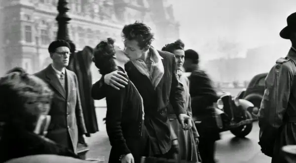 Pareja besándose apasionadamente en París, rodeada de gente y un coche antiguo. Un momento capturado en estas fotos históricas.