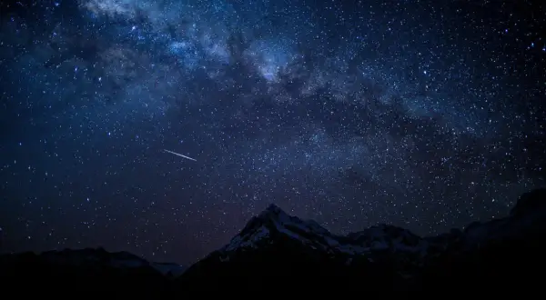 Una estrella fugaz cruza el cielo nocturno estrellado, con la Vía Láctea visible sobre montañas nevadas. Fotos perseidas capturan la magia.