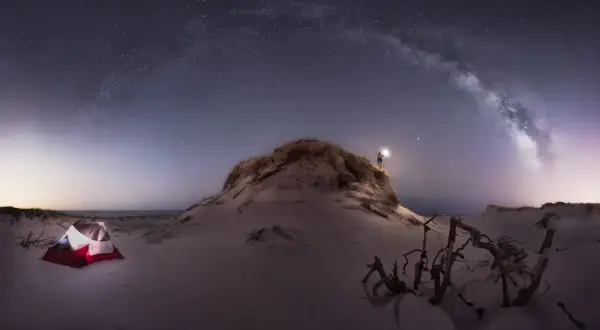Persona ilumina la noche estrellada con su linterna, cerca de una tienda de campaña en la playa. Las fotos con ruido capturan la magia del cielo nocturno.
