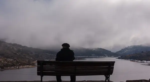 Hombre sentado en un banco, contemplando un lago y montañas cubiertas de niebla. La figura y el fondo crean una escena de introspección.