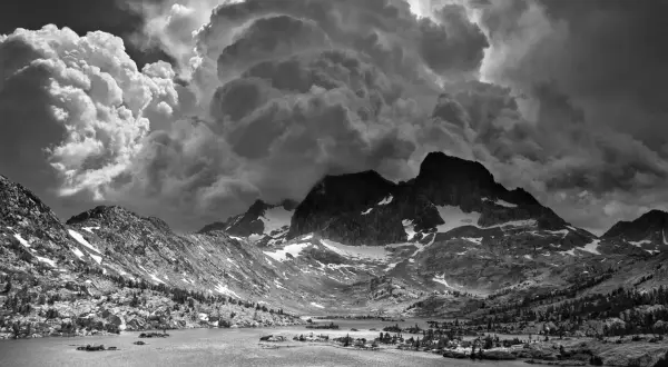 Paisaje montañoso dramático con nubes imponentes, típico de las ansel adams fotos. Un lago sereno refleja las cumbres nevadas.