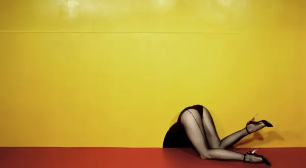 Piernas con medias de rejilla y tacones, estilo Guy Bourdin, asomando por un hueco contra un fondo amarillo y rojo.