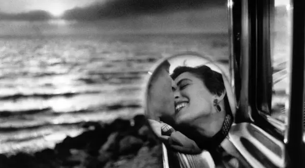 Una mujer sonríe en el espejo retrovisor de un coche, reflejando un beso. Elliott Erwitt capturó este momento íntimo junto al mar.