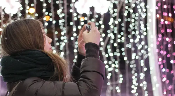 Chica con bufanda oscura toma foto de luces navideñas con un hermoso efecto bokeh.
