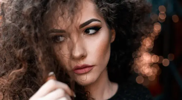 Plano medio corto de una mujer con cabello rizado y maquillaje dramático, mirando hacia un lado con una expresión pensativa.