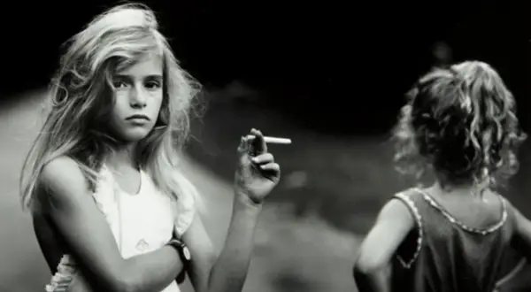 Una niña rubia, en una de las sally mann fotos, sostiene un cigarrillo. Otra niña, de espaldas, mira hacia la derecha.
