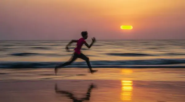 Un corredor se mueve velozmente en la playa al atardecer. El barrido fotográfico captura el movimiento y los colores cálidos del cielo y el mar.