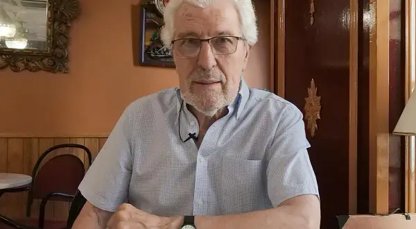 Miguel Soria, un hombre mayor con cabello blanco y gafas, sentado en un café con paredes naranjas y decoración clásica.