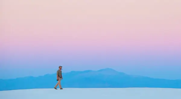 Hombre camina sobre dunas blancas bajo un cielo degradado de rosa y azul, respetando la ley del horizonte.