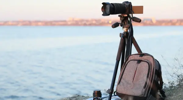 Equipo de fotografia basica listo para capturar el paisaje: cámara en trípode, mochila y lente junto al mar.