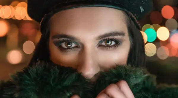 Ojos penetrantes de mujer con boina negra y abrigo de pelo verde, rodeada de luces bokeh. Un reto fotográfico nocturno.