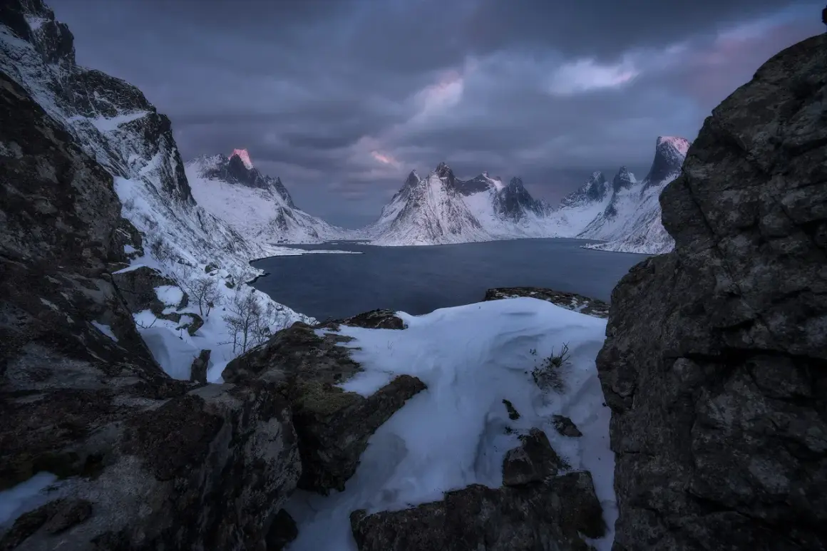 Foto panoramica de montañas nevadas y un lago oscuro bajo un cielo tormentoso. La luz del amanecer tiñe de rosa las cumbres.
