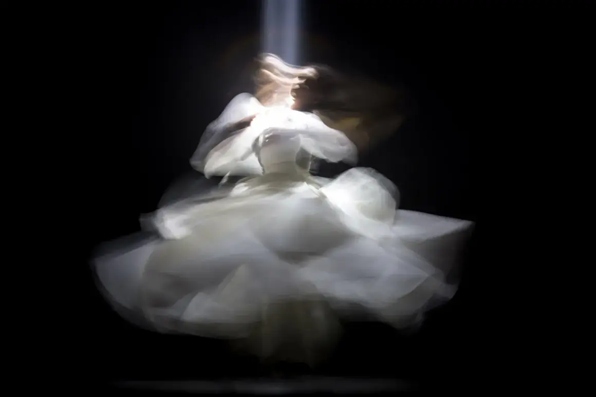 Figura etérea con un vestido blanco vaporoso, capturada con un **efecto foto movida** que evoca un baile onírico bajo un haz de luz.