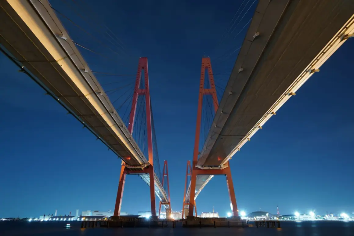 Dos torres rojas de un puente atirantado crean un efecto de fotos simétricas contra el cielo nocturno azul.